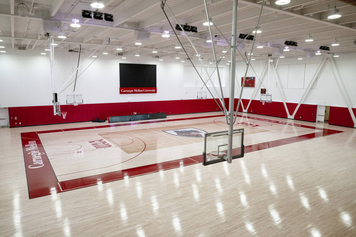 Large gym with a wood floor and athletics branding on it. Basketball hoops hand from the ceiling and a large screen hands on the wall.