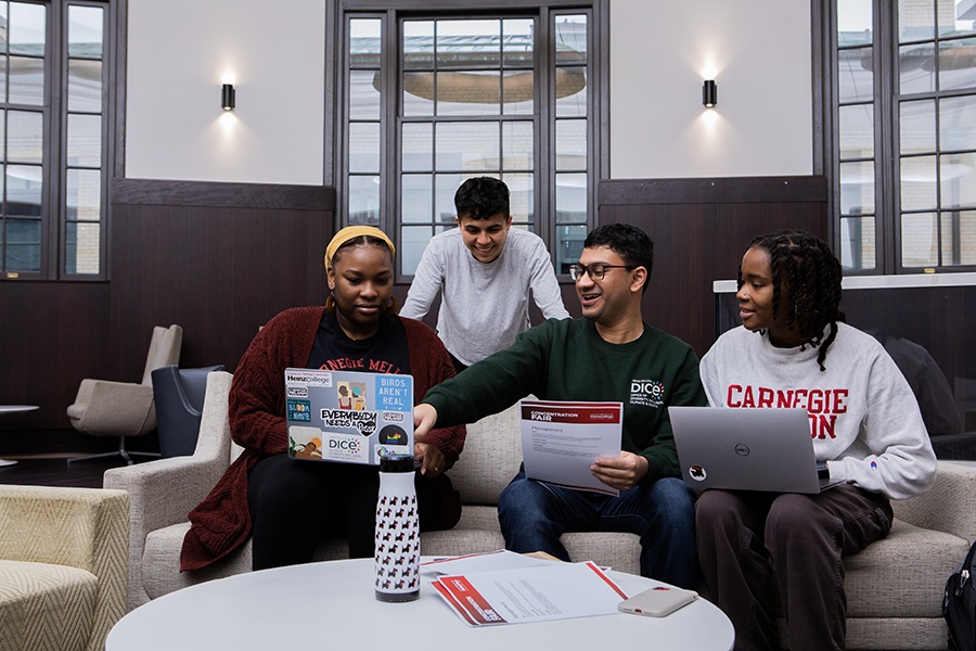 A group of four graduate students gathered in the Hamburg Hall Rotunda. Two women and one man are sitting, with the women holding laptops. The sitting man gestures to one laptop while holding a piece of paper. The standing man leans over to look at the screen.