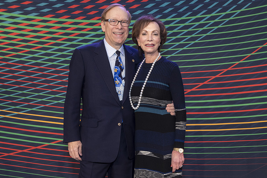 Tod Johnson stands in a dark suit with his arm around his wife Cindy in a dark dress with long pearl necklace.